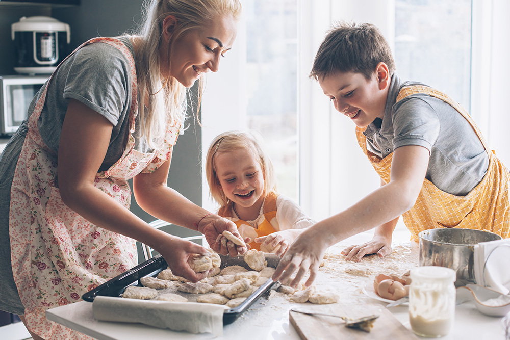 Beautiful blond mom teaching her two children cooking on the kitchen. Parent making everyday breakfast together with kids. Family at home lifestyle photo.